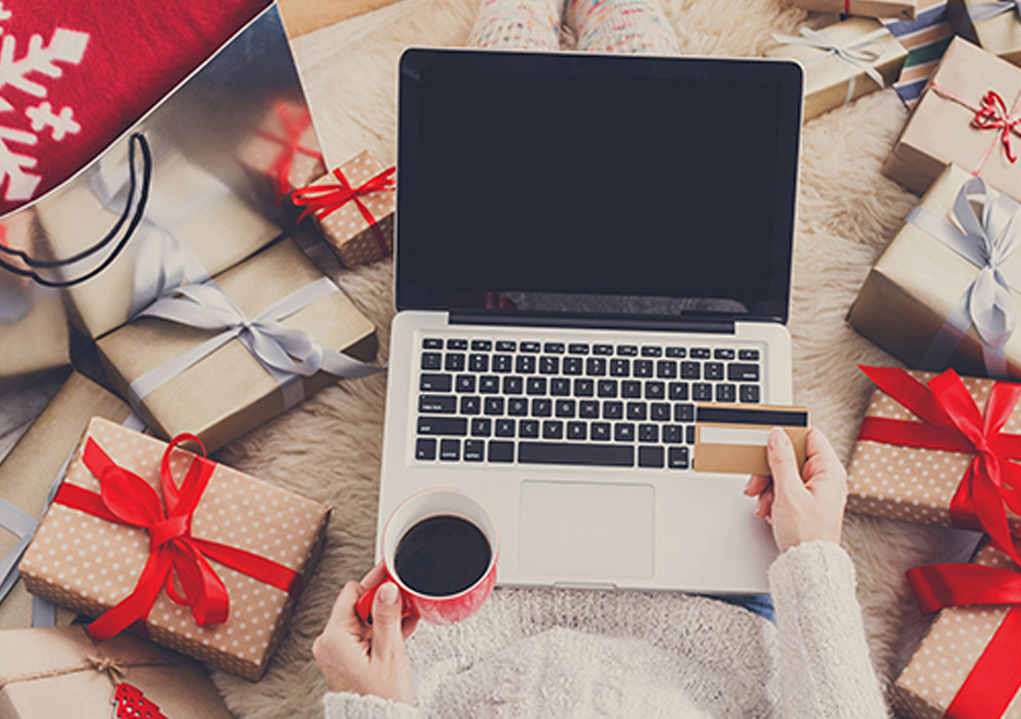 Woman sitting, surrounded by holiday presents, with laptop on her lap and shopping online